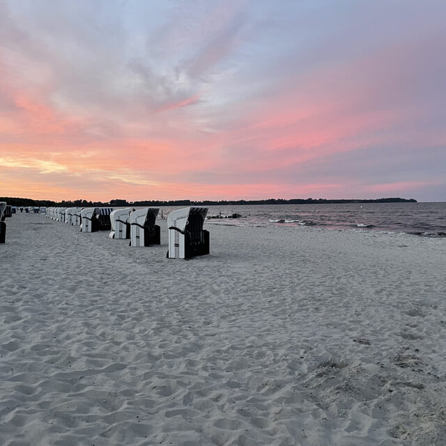Ostseestrand mit Strandkörben und Spaziergängern unter einem rosa gefärbten Abendhimmel.