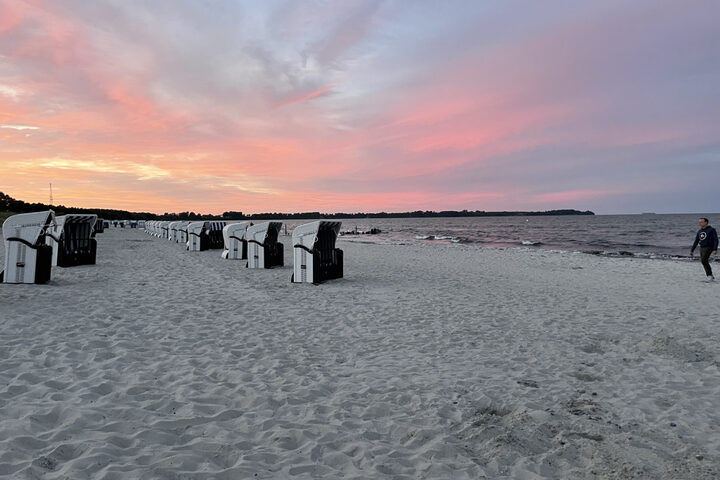 Strandkörbe und Spaziergänger am Ostseestrand unter einem rosa gefärbten Abendhimmel.
