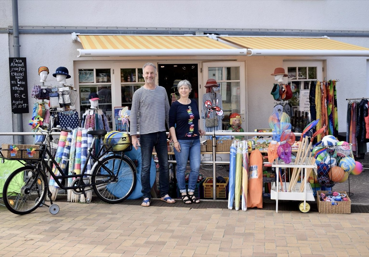 Ein Paar steht vor der Strandgut Buchhandlung in Juliusruh, daneben ein Fahrrad und bunte Strandartikel.