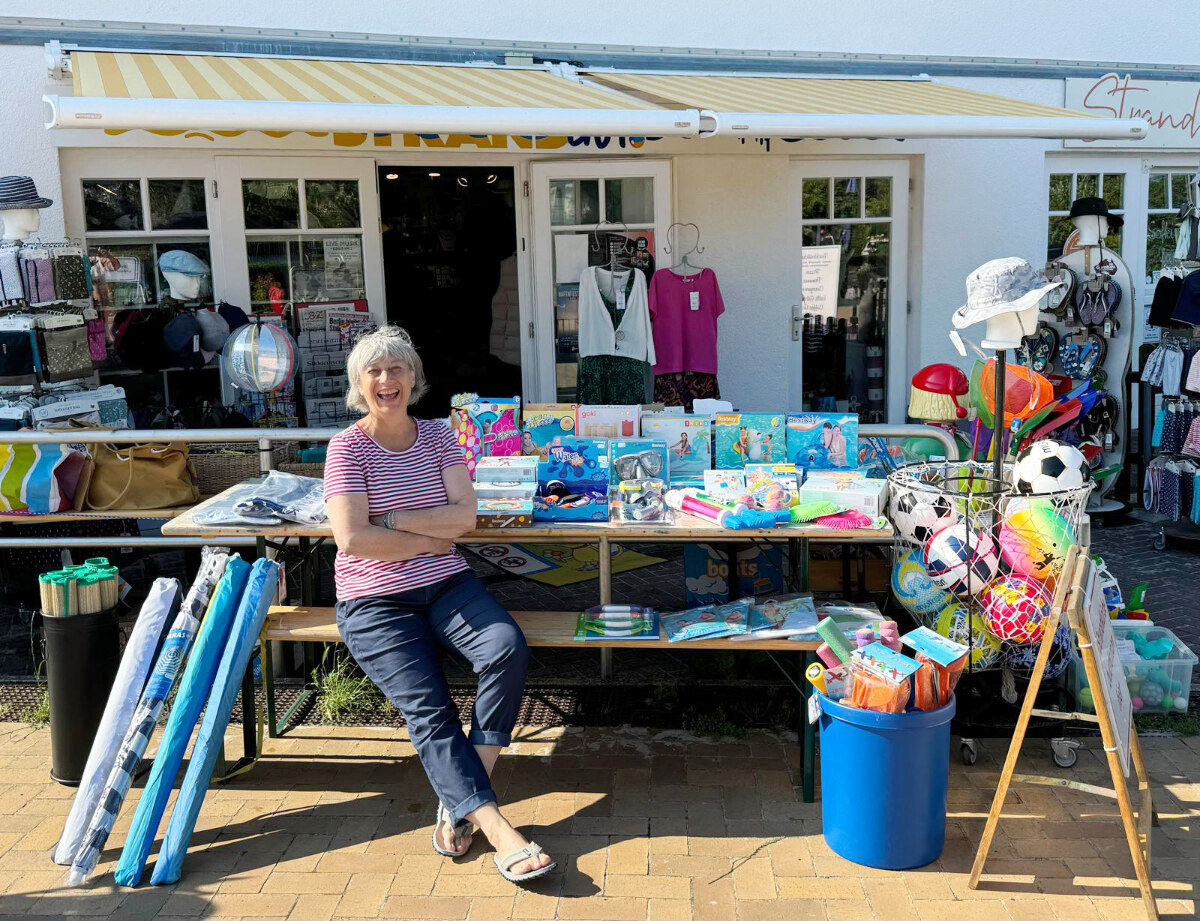Mitarbeiterin des Strandgut vor dem Laden, davor ein Tisch voller Strandspielzeug und Sommerartikel.